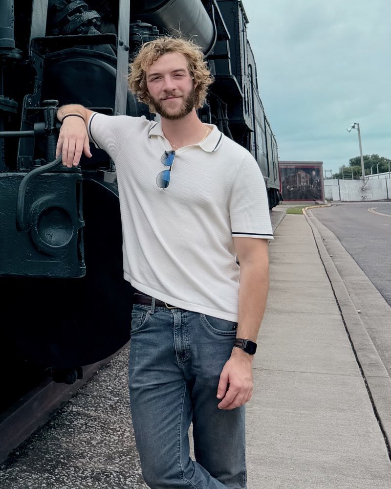 Benjamin Kotthoff in a white polo and jeans, leaning on a black steam locomotive